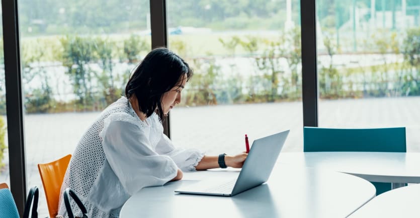 A woman in a white blouse sits with her laptop at a conference table positioned in front of floor-to-ceiling windows in a building on a college campus.