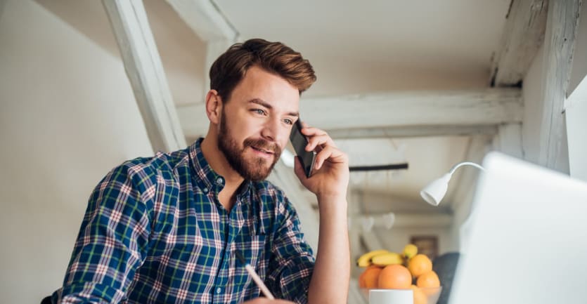 A man talks on a cell phone while gazing at a laptop and taking notes.