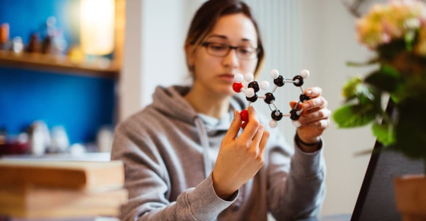 A young woman holds a molecule model in her hands while sitting at her computer.