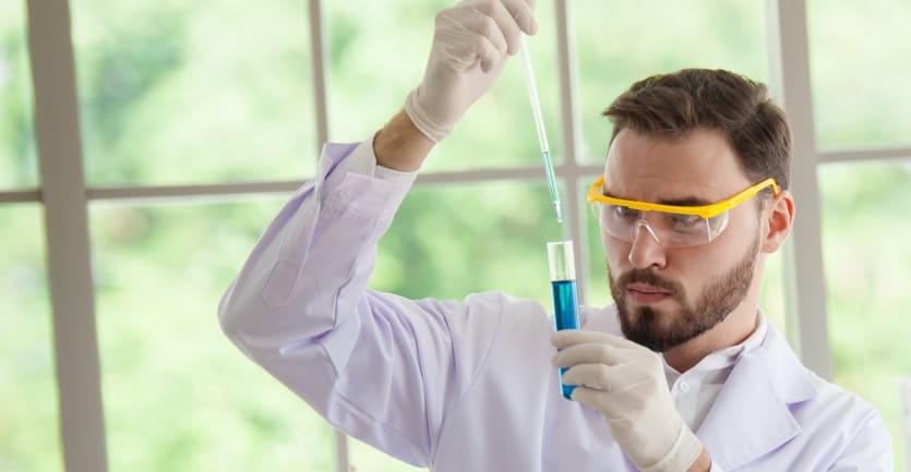 A scientist wearing gloves and safety glasses analyzes a blue liquid in a test tube.