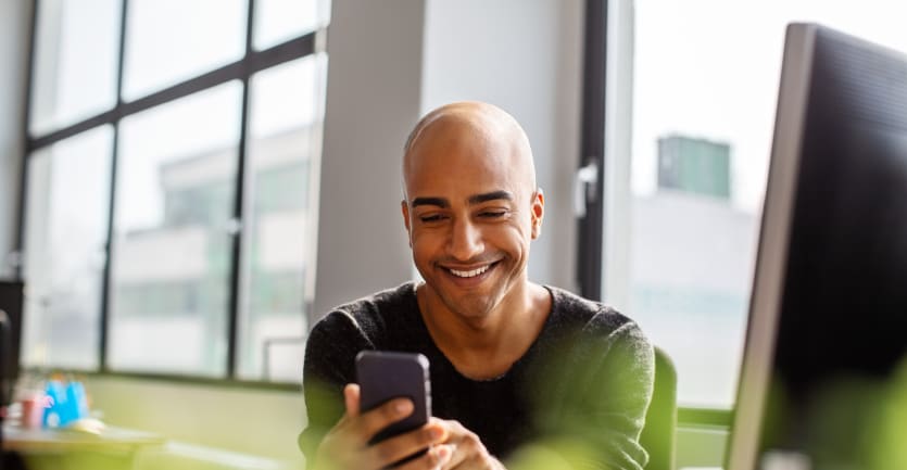 A young man happily checks his smart phone while sitting at his desk in a brightly lit office.