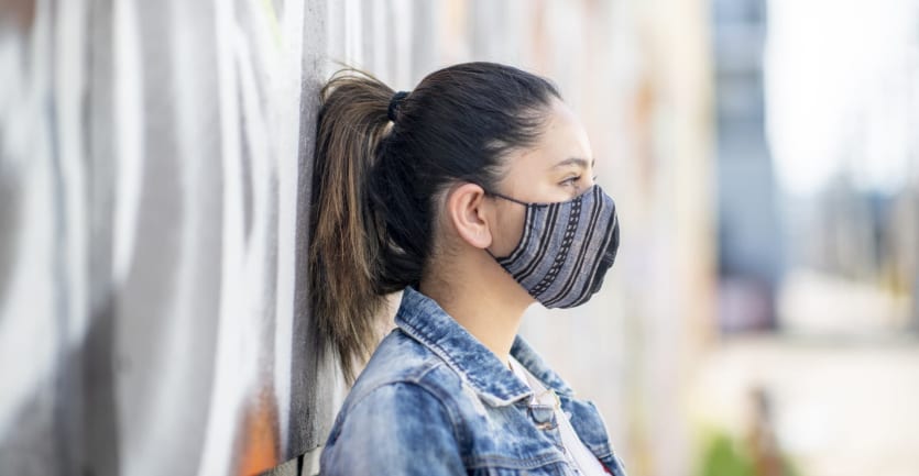 A college student wearing a jean jacket and face mask leans against a wall covered in graffiti.