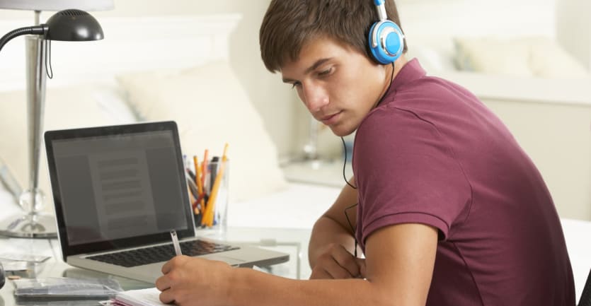 A teenage boy wearing a red T-shirt and blue headphones takes notes while studying in front of his laptop.