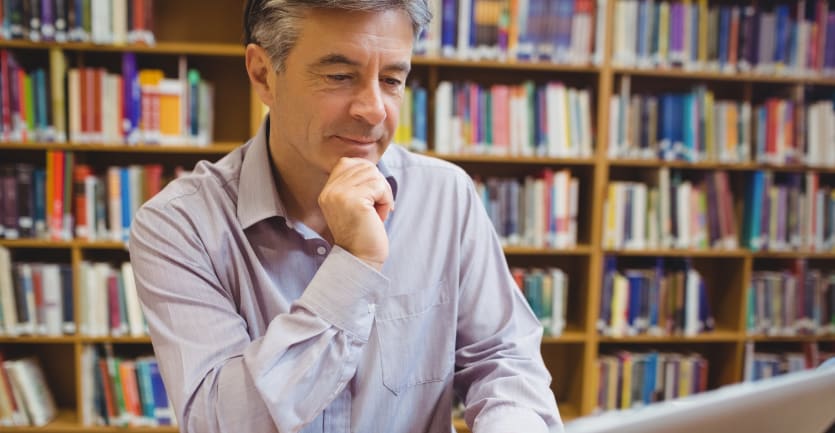 A middle aged man in a gray collared shirt answers emails on his laptop in an academic library.