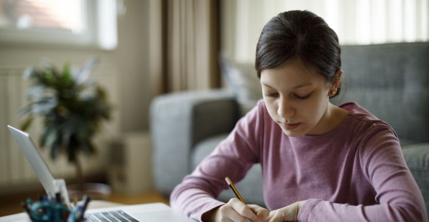 ピンクの長袖シャツを着た十代の少女がノートパソコンの前で勉強する。