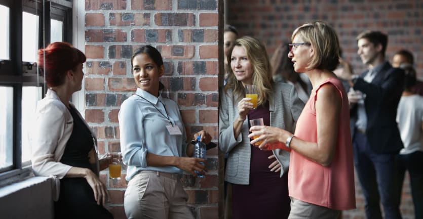 A group of smartly dressed women holding drinks chat while standing beneath a large window.