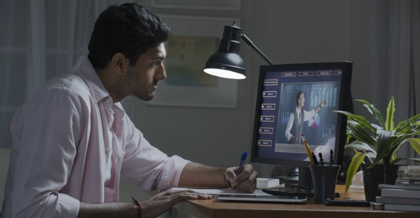 A man in an unbuttoned collard shirt with the sleeves rolled up takes notes as he follows along a professor's lecture on his laptop computer.