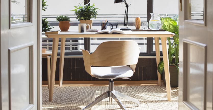 An empty chair and desk sit positioned before a large window; plants nestle in pots here and there, and an open book lies on the desk