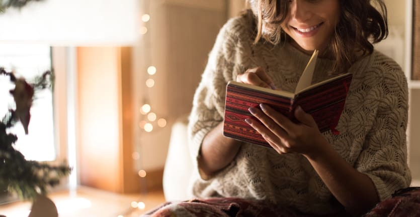 A young woman in a wool sweater sits cross-legged on the floor and smiles as she writes in a small journal