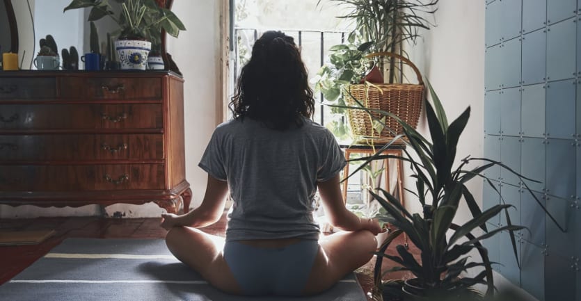 A woman sits in lotus position on the floor, surrounded by plants and facing an open window, her back turned toward the viewer