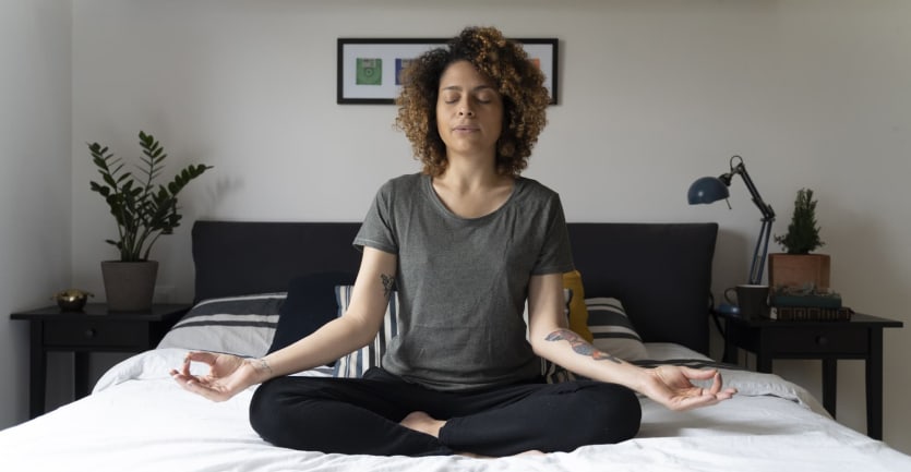 A woman sits cross-legged on her bed with her eyes closed deep in meditation.