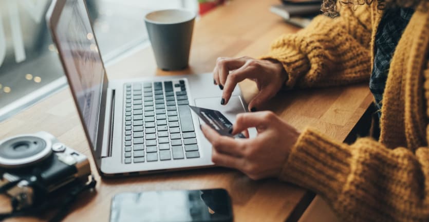 A woman wearing a comfortable cardigan holds out her credit card as she purchases an item online.