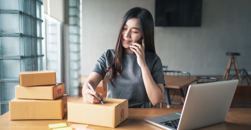 A woman talking on her smartphone writes down an addresss on a small cardboard box, ready to ship.