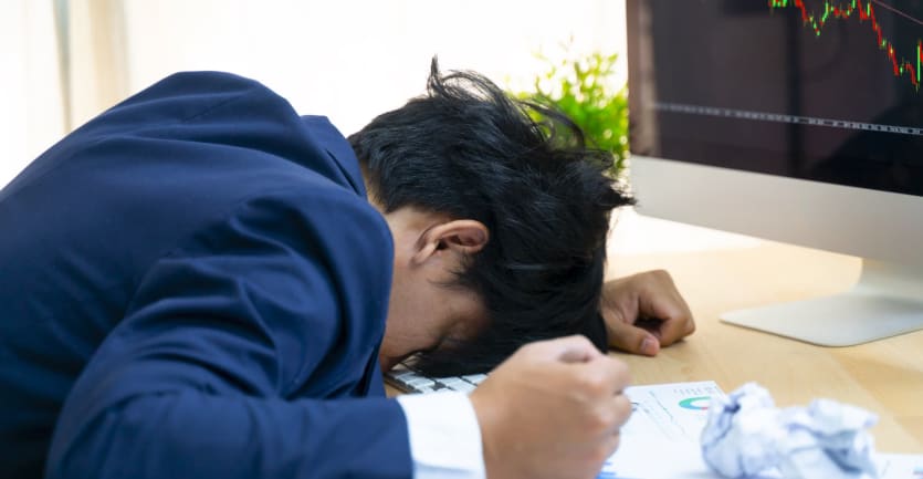 An anguished man in a suit clenches his fist at his desk as he lays his head down on his computer keyboard.