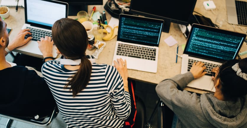 An overheaf shot of three developers consulting each others' laptops for an error in some code; their shared table is littered with a banana peel, coffee cups, and soda cans.