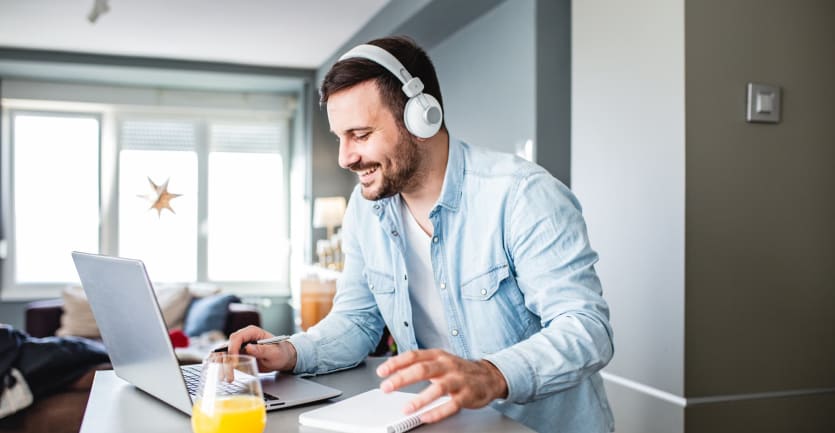 A man in a denim button-down shirt and over-ear headphones smiles as he looks at his laptop computer propped on his kitchen counter.