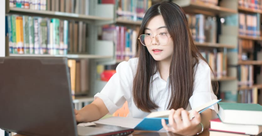 A woman in a short-sleeved blouse and glasses intently eyes her laptop computer with an open book in hand.