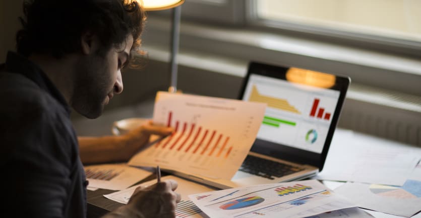 A man scribbles notes on printouts of graphs and charts sprawled across a busy desk.