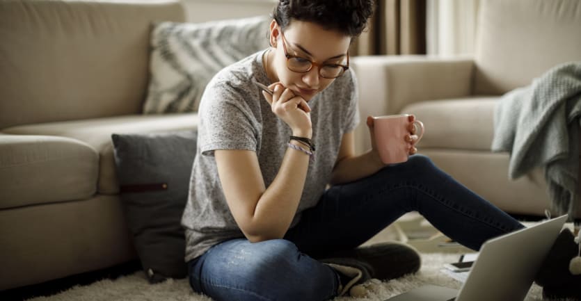 A young woman with short hair and glasses eyes her laptop computer as she sits on the floor of her living room.