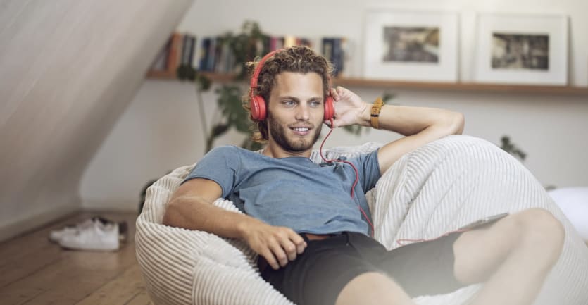 um homem de calções e t-shirt espreguiçando-se em casa, ouvindo um podcast nos seus fones de ouvido enquanto se espalhava num saco de feijão.