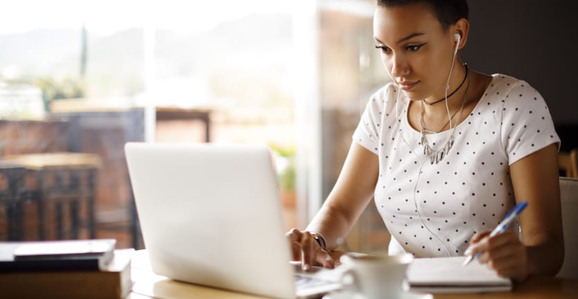 A young woman seated at a kitchen table reads from her laptop computer while taking handwritten notes/