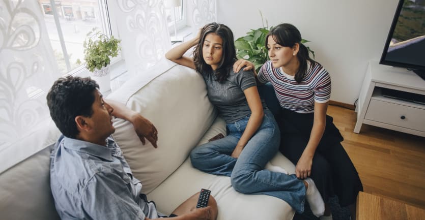 Two sisters sit on a living room couch and have a serious conversation with their father.