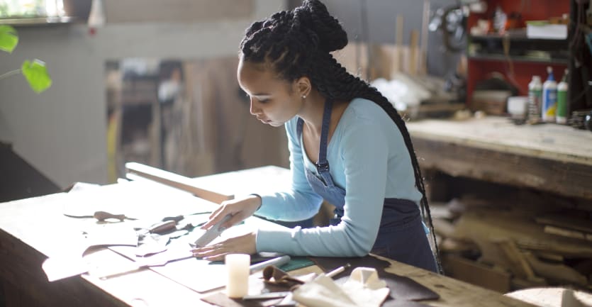 A young woman in long-sleeved t-shirt and apron carefully uses a utility knife to cut out materials on a large worktable.