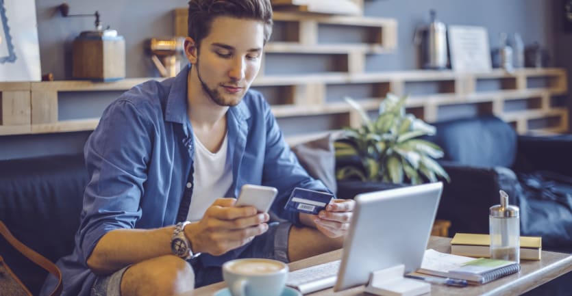 A young man in a sparse beard and jean shorts sits on a couch with an open laptop open on the coffee table before him, a credit card in one hand and his smartphone in the other.