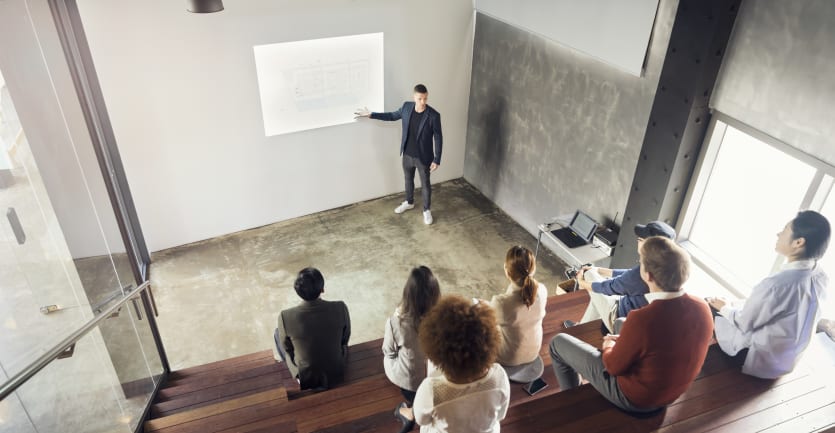 A man in a black t-shirt, blue sportscoat, jeans, and sneakers gives a presentation to a small group in a similarly small auditorium.