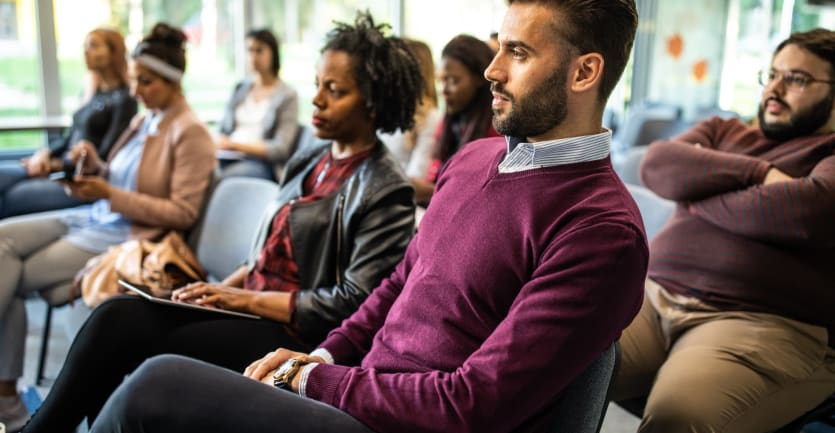 A diverse group of people in business-casual dress listen intently to a speaker in a small seminar.