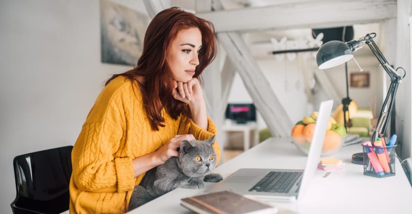 A young woman in an oversize wool sweater sits with her cat at her desk, scrolling through her laptop computer.