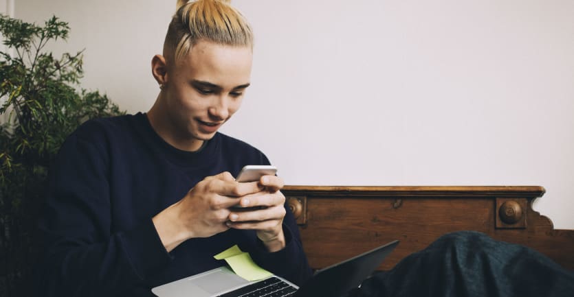 A young man with a top-knot in jeans and a sweatshirt checks his phone while he also conducts a sesrch on his laptop computer.