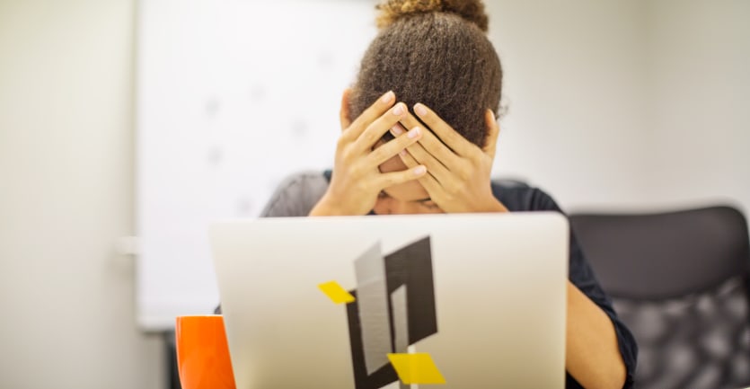 A woman sitting a table with her laptop computer holds her head in her hands in frustration.