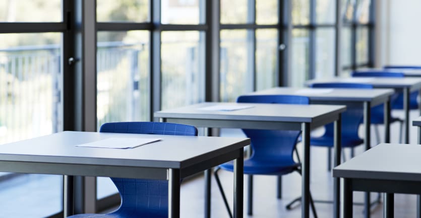 A row of desks in a classroom next to a large window sit empty.