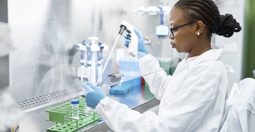 A woman in glasses, a labcoat, and latex gloves carefully handles chemical under a fume hood.