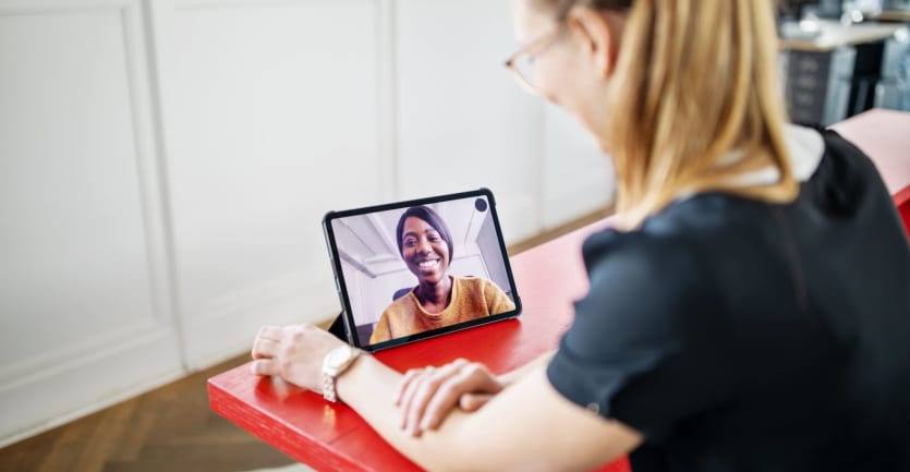 A young woman in a t-shirt and glasses holds a video call with a fellow student on her tablet computer.