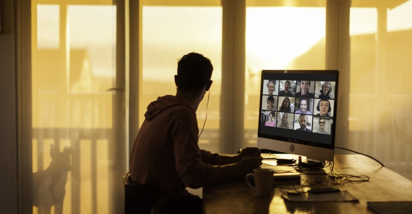 A man in a hoodie sweatshirt attends a virtual conference meeting on his desktop computer but turns his head to his balcony widnow, distracted by the silhouette of a dog.