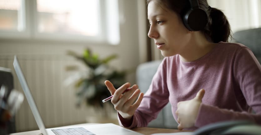 A young woman wearing headphones and a long-sleeved t-shirt sits at a dining table and holds a video-call on her laptop computer.