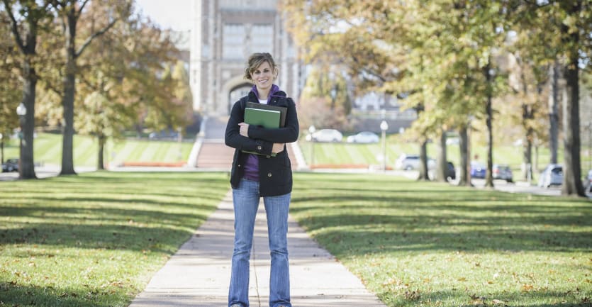 A woman in jeans, a hoodie, and an overcoat stands, smiling and holding several binders, in the middle of a college campus in the fall.