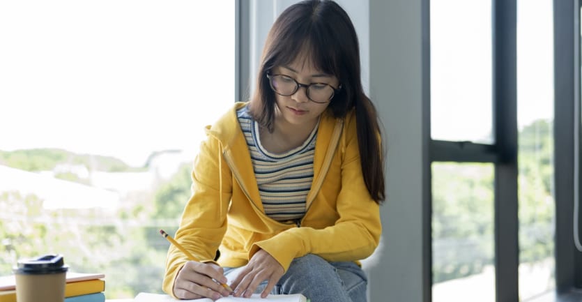 A woman in glasses and a brightly colored sweatshirt sits at a cafe table next to a floor-to-ceiling window and marks up a passage in a textbook.