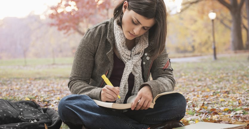 A woman wearing a jacket and scarf sits crosslegged on a foggy, leaf-strewn field on a college campus, highlighting a passage in a book.