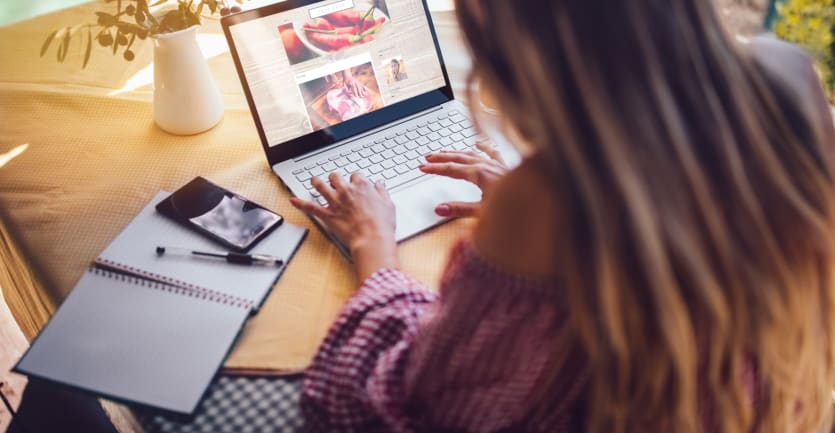 A woman looks over a food blog on her laptop computer set up on a dining table next to a pen, noteboook, smartphone, and vase of flowers.