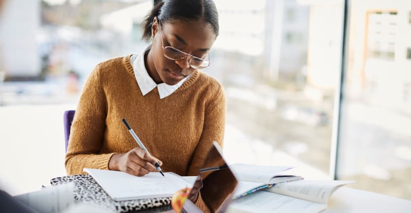 A young woman wearing glasses and a collared shirt underneath a sweater studiously writes in a notebook on na tabletop cluttered with papersa and laptop.