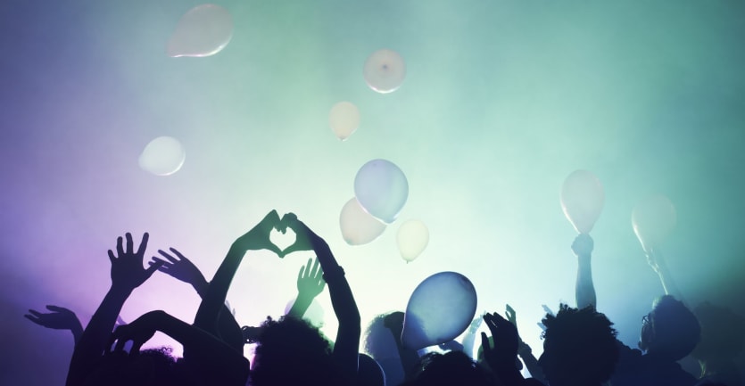 The silhouette of a crowd at a live concert is framed against smoke, lights, and balloons.