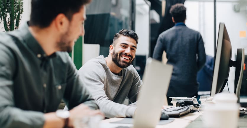 Two men in sharp, hip business casual wear smile and chat while seated at a row of desks in an open office.