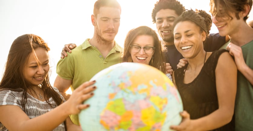 A multiethnic group of college students smile and hug as they touch and surround a globe.