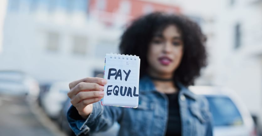 A woman in a dark t-shirt and denim jacket holds up a small reporter's notebook with the message PAY EQUAL scrawled in Sharpie.