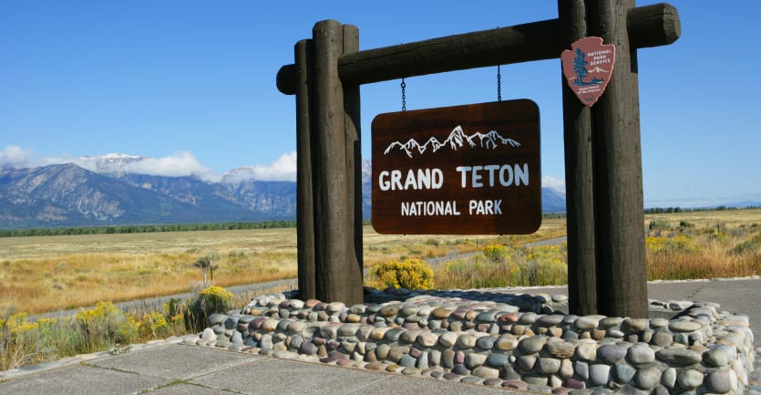 A large sign reading Grand Teton National Park stands against a serene backdrop of fields and distant mountains.