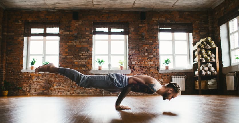 A shirtless man in sweatpants suspends himself from the floor using only his arms in a brightly lit yoga studio.