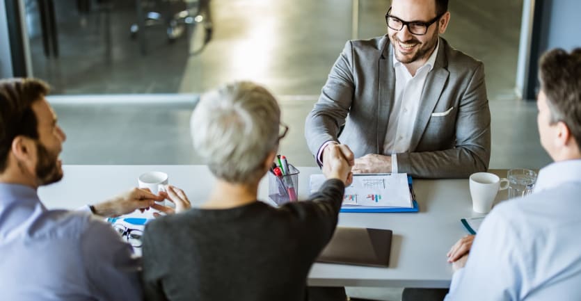 A smiling businessman wearing a suit and glasses shakes the hand of the businesswoman sitting across from him.